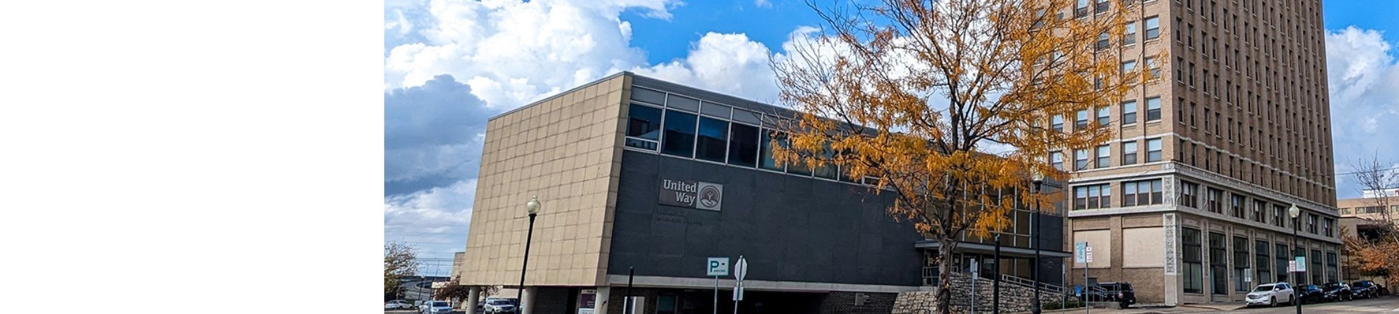 A modern United Way building with a slanted beige facade and large windows, located along a downtown street. A tree with autumn-colored leaves stands in front, and a tall historic-style building rises beside it under a partly cloudy sky.
