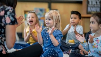 Teacher leading a group of young children clapping and engaging in a classroom activity, demonstrating positive interaction and early learning.