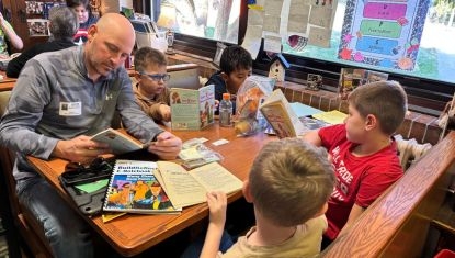 A volunteer reads aloud from a children's book while seated at a table with several elementary-age students. The children are gathered around, listening and following along with books in front of them. The group is participating in a reading activity inside a classroom or library setting, with colorful educational displays visible in the background. The scene highlights literacy, learning and positive adult engagement with children.